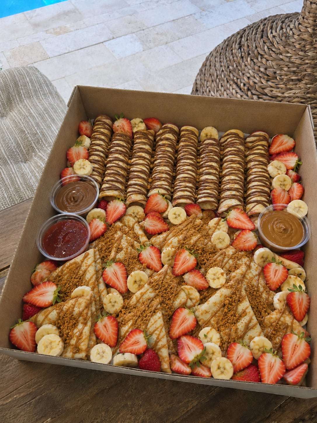 Open party box filled with rolled and folded crêpes, strawberries, bananas, sauces, and toppings